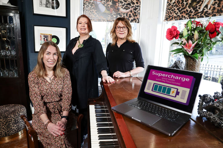 3 women around a grand piano smiling at the camera, with a laptop displaying the Supercharge logo.